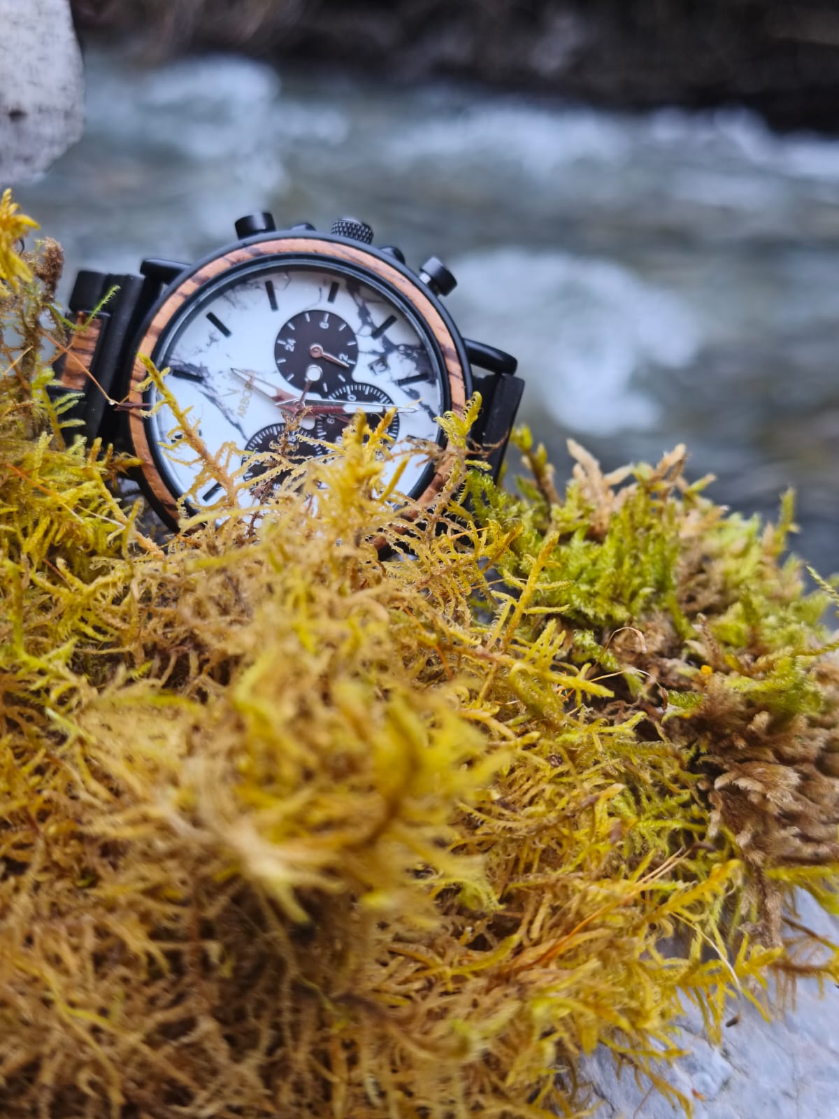 Montre en bois_Origine sur un tapis de mousse jaune a coté d'une rivière en pleine nature